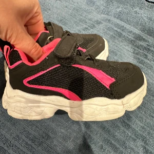 Svarta och rosa sneakers med kardborreband - Snygga och bekväma svarta sneakers med rosa detaljer och vit sula. Skorna har kardborreband för enkel på- och avtagning samt en sportig design. Perfekta för aktiva dagar.