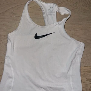 Vit linnetopp från Nike - Säljer en vit linnetopp från Nike med Dri-FIT-teknologi. Toppen har en sportig design med brottarrygg och ett svart Nike-logotyp på framsidan. Perfekt för träning och varma dagar.