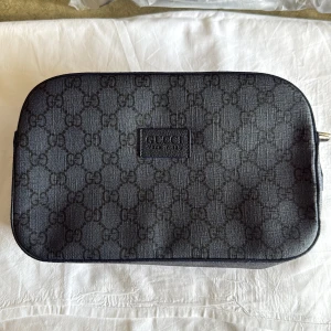 Svart axelväska från Gucci med GG-mönster - Snygg svart axelväska från Gucci med klassiskt GG-monogram över hela väskan. Väskan har en rektangulär form, dragkedja upptill och justerbar axelrem i textil och skinn. Perfekt för dig som vill ha en stilren och lyxig accessoar med ikonisk design.