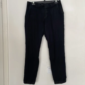 Svarta kostymbyxor från Dressmann - Svarta kostymbyxor från Dressmann i Super 100's ull. Klassisk rak modell med pressveck, sidfickor och bakfickor med knappar. Perfekta för en clean och stilren look. Byxorna har bälteshällor och stängs med knapp och dragkedja.