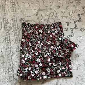 Blommig volangkjol från Talis - Supersöt kort kjol från Talis med blommigt mönster i rött, vitt, rosa och grönt. Kjolen har volangdetalj framtill och är i mjuk bomull. Perfekt för dig som vill ha en färgglad stil.