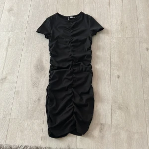 Svart kortklänning från H&M Divided - Svart kortklänning från H&M Divided med tight passform och rynkade detaljer längs hela klänningen. Klänningen har korta ärmar och rund halsringning. Perfekt för dig som gillar en stilren och trendig look.