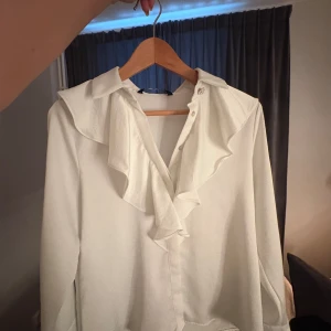 Vit blus med volangkrage - Snygg vit blus med stor volangkrage och knappar framtill. Långärmad modell med klassisk krage och dekorativa detaljer vid ärmsluten. Använt 2 gånger så den är i ny skick 