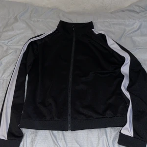 Svart vindjacka från Bik Bok med vita ränder - Sportig svart kofta från Bik Bok i storlek L. Kofta har vita ränder längs ärmarna, hög krage och hel dragkedja framtill. Tillverkad i ett glansigt syntetmaterial som ger en cool och avslappnad vibe. Perfekt för dig som gillar streetstyle.