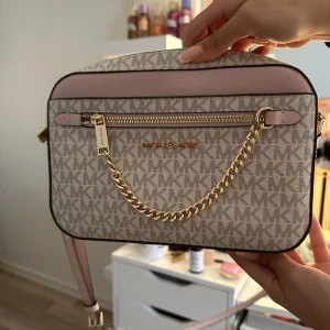 Michael Kors beige axelväska med kedja - Snygg beige axelväska från Michael Kors med MK-logga över hela väskan, ljusrosa detaljer och guldfärgad kedja framtill. Väskan har dragkedja och justerbar axelrem. Perfekt storlek för det viktigaste och riktigt trendig design.