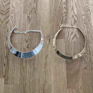 Chunky halsband i silver och guld - Två chunky halsband i metall, ett i blankt silver och ett i blankt guld. Båda har en bred, böjd form som ligger snyggt runt halsen och justerbar kedja i nacken. Perfekta statement-smycken för att lyfta din outfit.