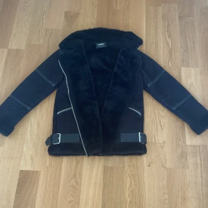 Svart pälsjacka från Pull&Bear - Snygg svart pälsjacka från Pull&Bear med fuskpäls på krage och ärmslut. Jackan har asymmetrisk dragkedja, två snedställda fickor med dragkedjor och coola detaljer i skinnimitation. Perfekt för kalla dagar när du vill se extra stylish ut.