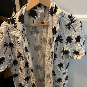 Vit skjorta med palmmönster Imperio Corinto - Snygg vit skjorta från Imperio Corinto med svarta palmer över hela tyget. Skjortan har korta ärmar, klassisk krage och knäppning framtill. Perfekt för sommarens varma dagar och ger en chill vibe. Tillverkad i Italien.