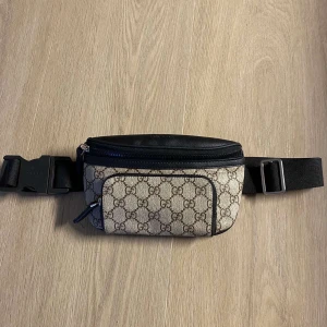 Gucci beige magväska med GG-mönster - Säljer en snygg magväska från Gucci i beige och svart med klassiskt GG-mönster. Väskan har justerbart band, flera fack med dragkedja och detaljer i svart skinn. Perfekt för att bära det viktigaste och ge outfiten en lyxig touch. ‼️Priset går att diskuteras‼️
