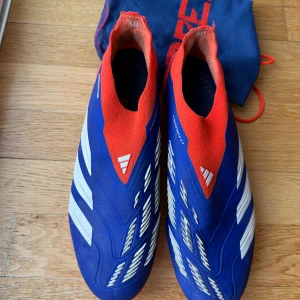 Adidas Predator Elite 43 1/3 - Säljer ett par Adidas Predator Elite fotbollsskor i blått och rött med vita detaljer. Storlek 43 och 1/3 Skorna har en strumpliknande passform utan snörning, mönstrad ovandel och tydliga Predator-loggor. Bra skick
