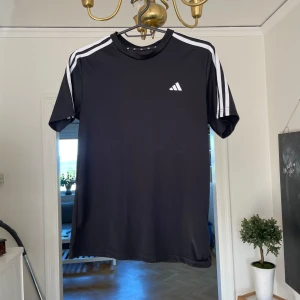 Adidas T-shirt - Svart t-shirt från Adidas med klassiska vita ränder längs axlarna och litet Adidas-logga på bröstet. Tillverkad i lätt och skönt polyestermaterial, perfekt för träning eller sport. Normal passform och korta ärmar.