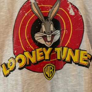 Looney Tunes t-shirt med Bugs Bunny - Gråmelerad t-shirt med stort tryck av Bugs Bunny och Looney Tunes-loggan i rött, gult och svart framtill. Klassisk passform med korta ärmar och rund hals. Perfekt för dig som älskar retro och tecknade serier. Den sitter som en L 
