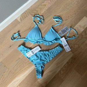 Ljusblå Julia bikini med volang - Supersöt ljusblå bikini från Gina trickot  med volangdetaljer. Triangeltopp med knyt i nacken och ryggen samt matchande bikiniunderdel med rynkade kanter. Perfekt för stranden eller poolen, i en fräsch himmelsblå färg. Säljs som set.