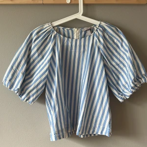 Blåvit randig blus från H&M - Superfin blus från H&M i blåvitt randigt bomullstyg. Modellen har puffiga korta ärmar med resår och rund halsringning. Perfekt för dig som gillar en luftig och trendig look med lite volym i ärmarna.