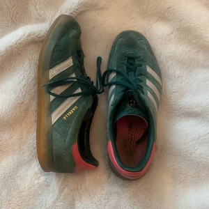Adidas Gazelle gröna mockasneakers - Säljer ett par Adidas Gazelle sneakers i grön mocka med klassiska vita ränder och gummisula. Skorna har snörning, detaljer i rosa vid hälen och guldfärgad Gazelle-text på sidan. 