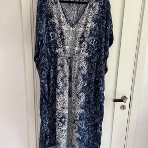 Klänning från Indiska - Snygg långklänning från Indiska i marinblått och vitt med paisleyinspirerat mönster. Klänningen har v-ringning, lös passform och knytband i midjan. Perfekt för dig som gillar bohemisk stil och vill ha något luftigt och bekvämt.