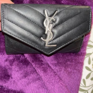Svart YSL plånbok i skinn - Snygg svart plånbok från YSL i äkta skinn med silverfärgad logga framtill. Plånboken har quiltad design med struktur och flera fack för kort och sedlar. Perfekt accessoar för dig som gillar lyxiga detaljer och stilren look.