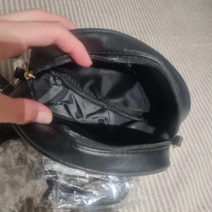 Svart axelväska med tofs från SHEIN - Snygg svart axelväska från SHEIN i rundad form med gulddetaljer och dekorativ tofs. Väskan har dragkedja upptill, ett mindre fack framtill och justerbar axelrem. Tillverkad i syntetmaterial med slät finish och stilren look.