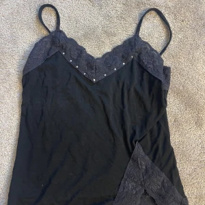 Svart linnetopp med spets från Effgee - Svart linnetopp från Effgee med tunna axelband och v-ringning. Toppen har dekorativ spets både upptill och nertill samt små silvriga pärlor längs urringningen. Perfekt för dig som gillar en stilren men ändå detaljrik look.