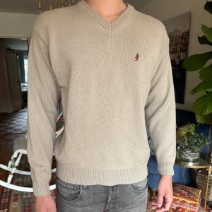 Beige stickad tröja Marlboro Classics - Beige stickad tröja från Marlboro Classics med v-ringad hals och diskret broderad logga på bröstet. Tröjan har ribbade muddar och är långärmad, perfekt för lager-på-lager under kyliga dagar. Enkel och stilren design som funkar till det mesta.