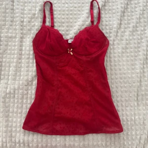 Röd spetsig korsett med rosett - Supersnygg röd korsett från Intimissimi med tunna axelband, spetsdetaljer och liten rosett framtill. Figurnära passform och lätt transparent tyg med diskret mönster. Perfekt att styla med jeans eller kjol för en trendig look.