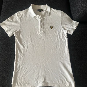 Vit pikétröja från Lyle & Scott - Klassisk vit pikétröja från Lyle & Scott med korta ärmar och broderad gul logga på bröstet. Tröjan har krage och knäppning framtill. Perfekt för en clean och stilren look.