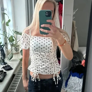 Vit prickig off shoulder topp med snörning - Supersöt vit topp med svarta prickar och puffiga korta ärmar. Toppen har snörning på sidorna och knytband nertill för en extra snygg detalj. Offshoulder-modell med smala axelband och figurnära passform. Perfekt för en trendig och lekfull look.