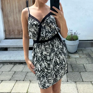 Svartvit omlottklänning med mönster - Snygg kort klänning med svartvitt grafiskt mönster och omlott-skärning. Klänningen har smala axelband, markerad midja och svarta detaljer längs kanterna. Perfekt för dig som gillar en cool och modern stil med lite edge.