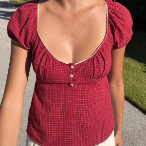 Rutig röd blus med puffärm - Supersöt röd blus med små vita rutor, puffiga korta ärmar och djup v-ringning med spetskant. Tre knappar framtill ger en vintagevibe. Perfekt till sommarens alla outfits och lätt att matcha med jeans eller kjol.
