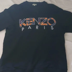 Svart Kenzo sweatshirt med tryck - Svart sweatshirt från Kenzo med stort broderat Kenzo Paris-tryck i orange, blått och vitt på bröstet. Klassisk rund halsringning och lång ärm. Perfekt för dig som gillar streetstyle och vill sticka ut med en ikonisk logga. Går att diskutera pris
