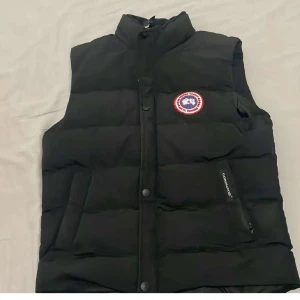 Svart dunväst från Canada Goose - Snygg svart dunväst från Canada Goose med hög krage och klassisk logga på bröstet. Västen har dragkedja och knappar framtill samt två fickor med dragkedja. Perfekt för lager-på-lager och håller dig varm under kyliga dagar.