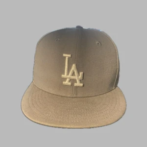Grå LA keps från New Era - Snygg grå keps från New Era med LA-logga broderad framtill och MLB-logga bak. Klassisk flat brim och stilren look som passar till avslappnad stil. Kepsen är tillverkad i bomull och har en neutral färg som är lätt att matcha. Köparen står för frakt.