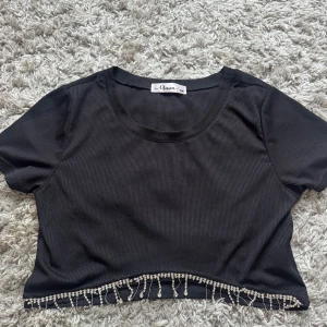 Svart croppad topp med pärldetaljer - Svart ribbad croppad topp från Unica med korta ärmar och rund halsringning. Nedtill finns snygga pärldetaljer som hänger och ger toppen en extra touch. Perfekt för dig som vill sticka ut lite extra med din outfit.