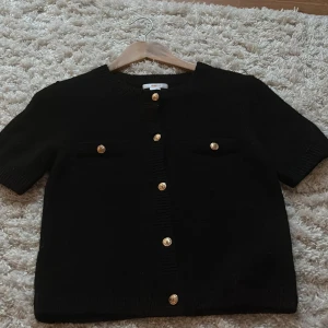 Svart kortärmad stickat kofta - Svart kortärmad stickad kofta med guld knappar. Den är ganska liten i storleken, jag brukar ha storlek 158 i ginas andra tröjor men i denna behövde jag gå upp till 170 för att den annars var för kort! Aldrig använt! 