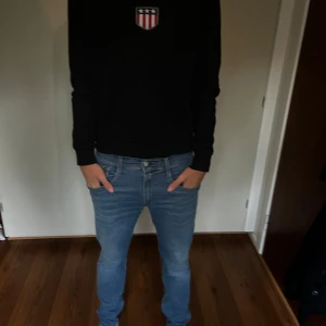 Svart sweatshirt från GANT - Svart sweatshirt från GANT med broderad logga och sköld på bröstet. Tröjan har rund halsringning och långa ärmar. Klassisk och stilren design med vita och röda detaljer i loggan. Perfekt för en avslappnad och snygg look.