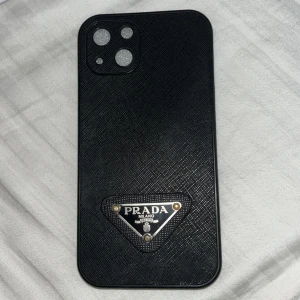 Svart Prada mobilskal i skinnlook - Snyggt svart mobilskal från Prada med klassisk triangel-logga i metall. Skalet har en lyxig skinnstruktur och insidan är beige med präglad text och dekorativa detaljer. Perfekt accessoar för dig som vill ha en exklusiv vibe på mobilen.