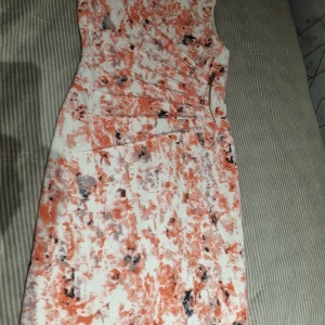 Kort klänning med orange mönster - Snygg kort klänning med abstrakt mönster i orange, vitt och svarta detaljer. Figurnära passform och dragkedja i sidan. Perfekt för dig som vill sticka ut med färg och stil.