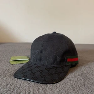 Svart Gucci keps - Snygg svart keps från Gucci med klassiskt GG-monogram över hela kepsen. Kepsen har en böjd skärm och ikoniska gröna och röda ränder på sidan. Tillverkad i bomull med justerbar rem baktill för perfekt passform.