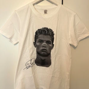 Vit t-shirt med Cristiano Ronaldo  - Vit t-shirt i 100% bomull med svartvit porträtt-tryck på Ronaldo framtill och signatur, samt stor grå logga och texten 'CUZ ANYTHING IZ YOURS' på ryggen. Klassisk passform och rund hals. Perfekt för dig som gillar statement-plagg. Plagget är helt nytt men på grund av frakten så har den blivit skrynklig.
