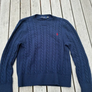 Mörkblå kabelstickad tröja Polo Ralph Lauren - Kabelstickad mörkblå tröja från Polo Ralph Lauren i ull, med klassisk rund hals och röd broderad logga på bröstet. Långärmad modell med ribbade muddar och nederkant. Perfekt för dig som gillar preppy och tidlös stil.