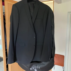 Svart kostym från Tiger of Sweden - Klassisk svart kostym från Tiger of Sweden i stilren design. Kavaj med två knappar, smala slag och matchande byxor med pressveck. Kostymen är tillverkad i ullblandning och har en elegant, tidlös look som passar perfekt till mer uppklädda tillfällen. Kostym storlek 152, kostymbyxa c154, väst storlek 56