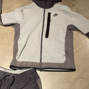 Nike tech, storlek M men passar bra som S - Nike vindjacka,byxor i ljusgrått och mörkgrått med hel dragkedja, huva och svart logga på bröstet. Jackan har en snygg kontrastdesign, lång ärm och en vertikal bröstficka med dragkedja. Perfekt för dig som gillar sportig och clean stil.