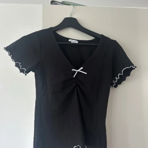 Svart topp med rosett och volang - Supersöt svart topp från Brandy Melville med v-ringning, liten vit rosett framtill och volangkantade ärmar och nederdel. T-shirten har korta ärmar och kontrastsömmar i vitt som ger en extra detalj.