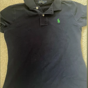 Marinblå pikétröja Polo Ralph Lauren - Klassisk marinblå pikétröja från Polo Ralph Lauren med grönt broderat logotyp på bröstet. Tröjan har korta ärmar, krage och två knappar framtill. Tillverkad i bomull för en bekväm och stilren look. Aldrig använt helt ny.