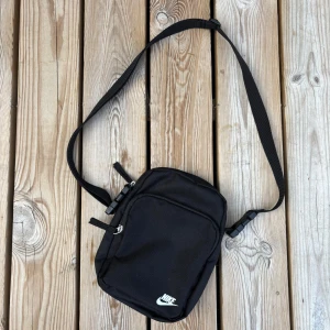 Svart axelväska från Nike - En stilren svart axelväska från Nike med justerbar rem och två fack med dragkedja. Väskan är tillverkad i slitstark polyester och har en diskret Nike-logga framtill. Perfekt storlek för mobil, plånbok och andra småprylar.
