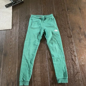 Gröna mjukisbyxor från The North Face - Snygga gröna mjukisbyxor från The North Face med resår och dragsko i midjan. Byxorna har ribbade muddar vid bensluten, två sidofickor och en vit logga på låret. Perfekta för chill eller träning.