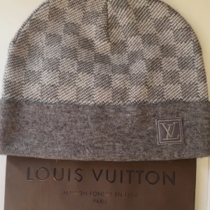Grå rutig mössa från Louis V - Exklusiv grå mössa  med rutigt mönster och logga framtill. Tillverkad i mjukt material som känns lyxigt mot huden. Perfekt accessoar för dig som vill sticka ut med stilren design och ikonisk branding.