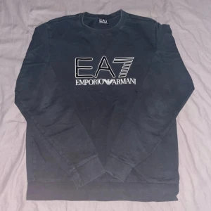 Svart EA7 Emporio Armani sweatshirt - Svart sweatshirt från EA7 Emporio Armani med stort logotryck i vitt och holografiska detaljer på bröstet. Klassisk rund halsringning och långa ärmar. Tillverkad i mjuk bomull för en skön och avslappnad stil.