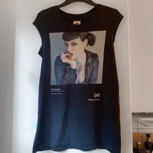 Linne från Lee - Svart ärmlös topp från Lee med ett stort tryck framtill som visar en person med tatuering och skinnjacka. Rund halsringning och rak passform. Materialet är mjuk bomull som känns skönt mot huden. Några små hål som syns i bilden. Meddela gärna för mått eller andra frågor☺️