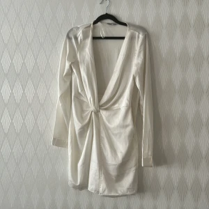 Vit omlottklänning med knut - Snygg vit kortklänning i satinliknande material med lång ärm och djup v-ringning. Klänningen har en omlott-design med knut framtill och dragkedja i sidan för enkel påklädning. Perfekt för dig som gillar stilrena och trendiga plagg.